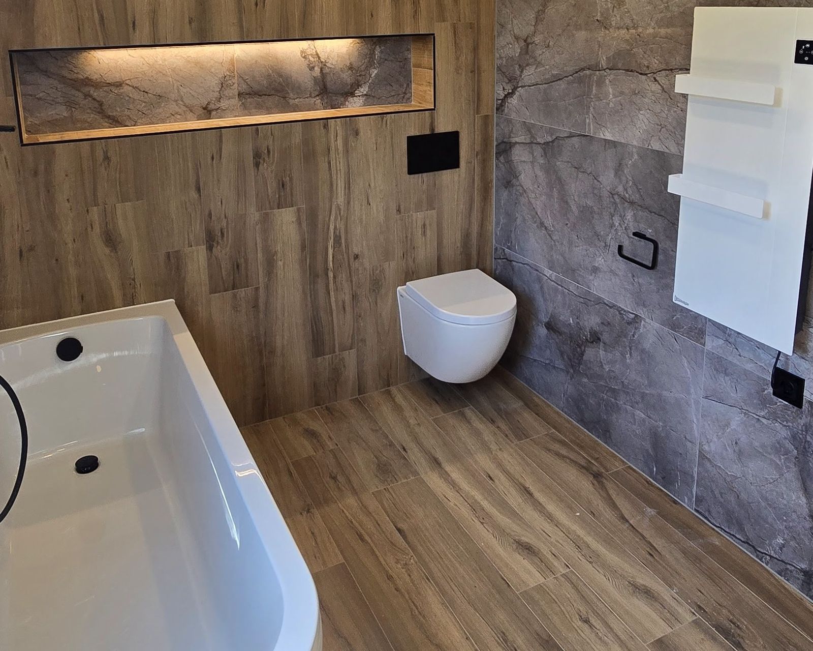 Badkamer met vrijstaand bad, houtlook vloer en wand met verlichte nis, hangend toilet en grijze marmerlook tegels