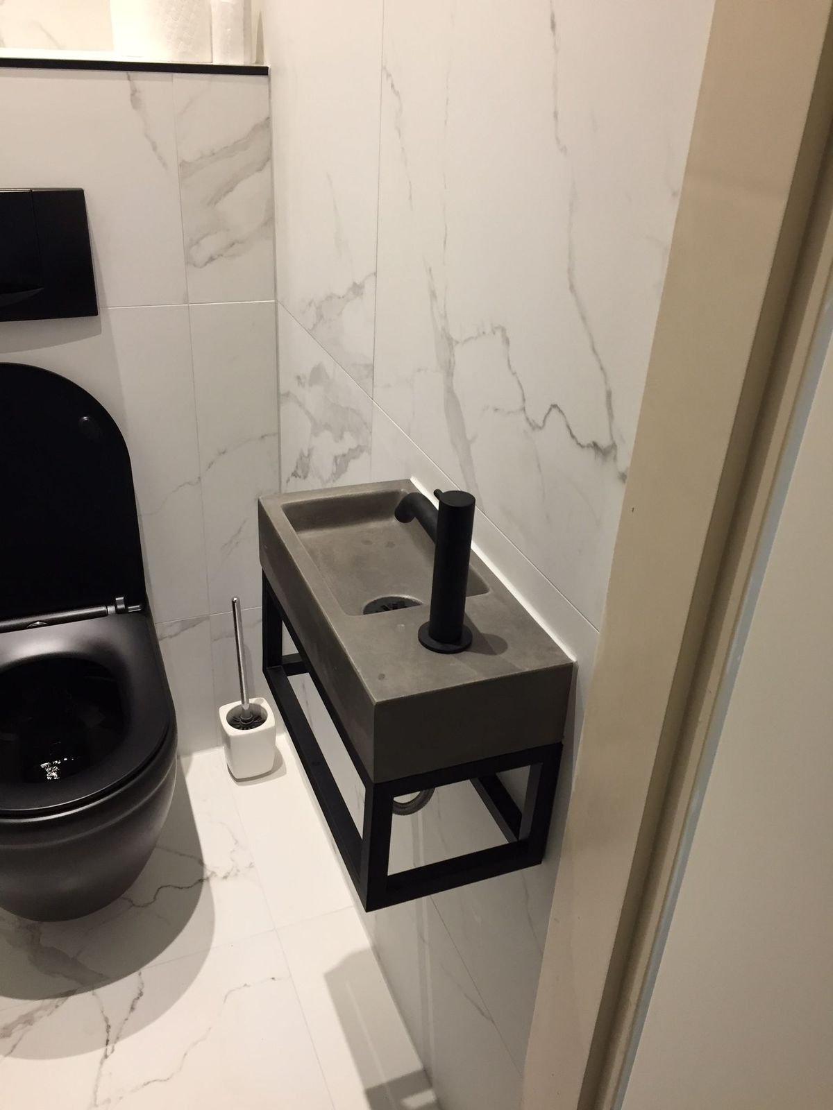 Toiletruimte met witte marmerlook tegels, zwart hangend toilet en een betonlook fonteintje op een zwart frame