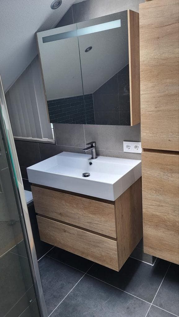 Witte wastafel op een houtlook meubel met spiegelkast onder een schuin plafond