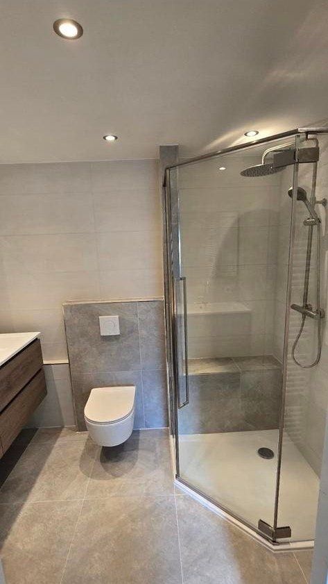 Glazen hoekdouche naast een hangend toilet in lichtgrijze tegels